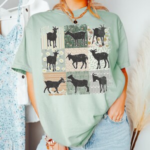 May include: A light green t-shirt with a patchwork design featuring nine black silhouettes of goats. Each goat is set against a different floral patterned background.
