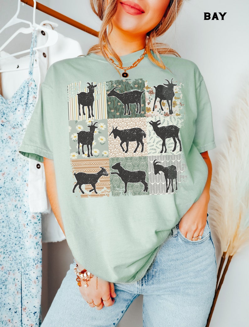 May include: A light green t-shirt with a patchwork design featuring nine black silhouettes of goats. Each goat is set against a different floral patterned background.