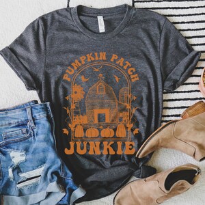 Puede incluir: Una camiseta gris oscuro con un dise&ntilde;o gr&aacute;fico naranja de un huerto de calabazas con un granero, calabazas y el texto "Pumpkin Patch Junkie".