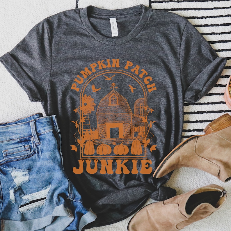 Puede incluir: Una camiseta gris oscuro con un dise&ntilde;o gr&aacute;fico naranja de un huerto de calabazas con un granero, calabazas y el texto "Pumpkin Patch Junkie".