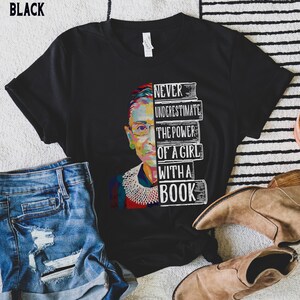Op de afbeelding: Zwart t-shirt met een kleurrijke afbeelding van een vrouwengezicht en de tekst "NEVER UNDERESTIMATE THE POWER OF A GIRL WITH A BOOK". Het ontwerp bevat een stapel boeken.