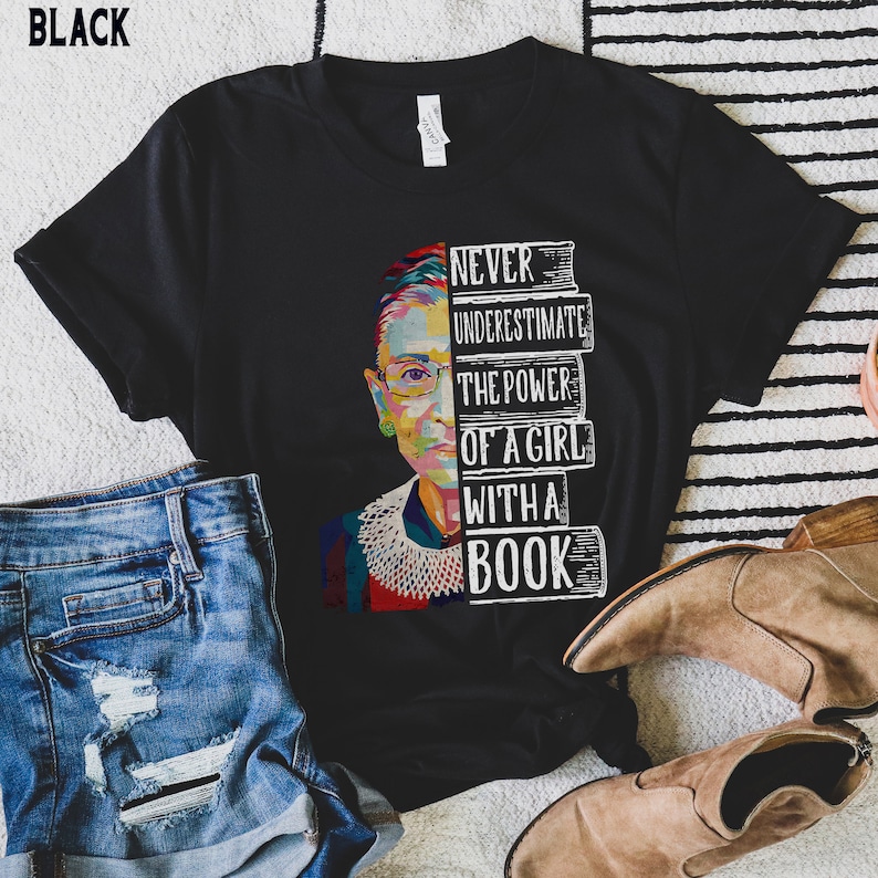 Op de afbeelding: Zwart t-shirt met een kleurrijke afbeelding van een vrouwengezicht en de tekst "NEVER UNDERESTIMATE THE POWER OF A GIRL WITH A BOOK". Het ontwerp bevat een stapel boeken.