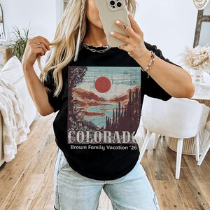 May include: Black t-shirt featuring a Colorado landscape design with a red sun, mountains, and trees. The text "COLORADO" and "Brown Family Vacation '26" are printed below the graphic. The shirt is a classic crew neck.