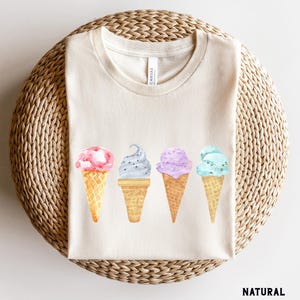 May include: A white t-shirt with a graphic of four ice cream cones in different colors and flavors. The ice cream cones are arranged in a row from left to right. The ice cream flavors are pink, gray, purple, and blue.