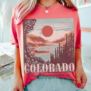 May include: Watermelon-colored t-shirt featuring a Colorado landscape design with a sun, mountains, and a river. The word "COLORADO" is printed in large letters, with "Olivia's Bachelorette '26" below. The shirt is a short-sleeved style.