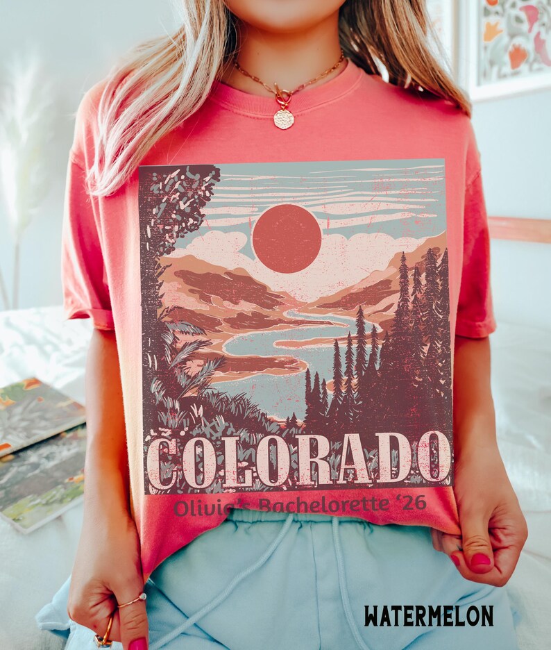 May include: Watermelon-colored t-shirt featuring a Colorado landscape design with a sun, mountains, and a river. The word "COLORADO" is printed in large letters, with "Olivia's Bachelorette '26" below. The shirt is a short-sleeved style.