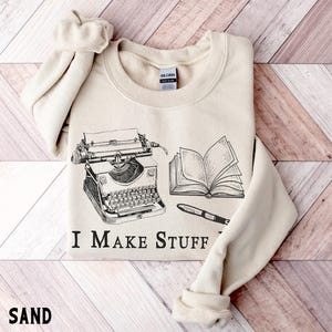 Puede incluir: Sudadera color arena con un gr&aacute;fico negro de una m&aacute;quina de escribir, un libro abierto y un bol&iacute;grafo. El texto "I MAKE STUFF" est&aacute; impreso debajo. La palabra "SAND" est&aacute; impresa en la parte inferior.