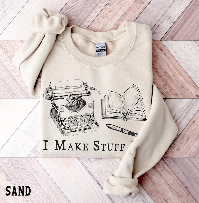 Puede incluir: Sudadera color arena con un gr&aacute;fico negro de una m&aacute;quina de escribir, un libro abierto y un bol&iacute;grafo. El texto "I MAKE STUFF" est&aacute; impreso debajo. La palabra "SAND" est&aacute; impresa en la parte inferior.