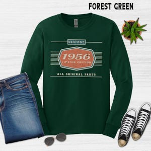 May include: Forest green long-sleeve shirt with a vintage design. The design includes the year "1956" in an orange and white hexagon, with the text "Limited Edition" and "All Original Parts". The shirt is paired with blue jeans and black and white sneakers.