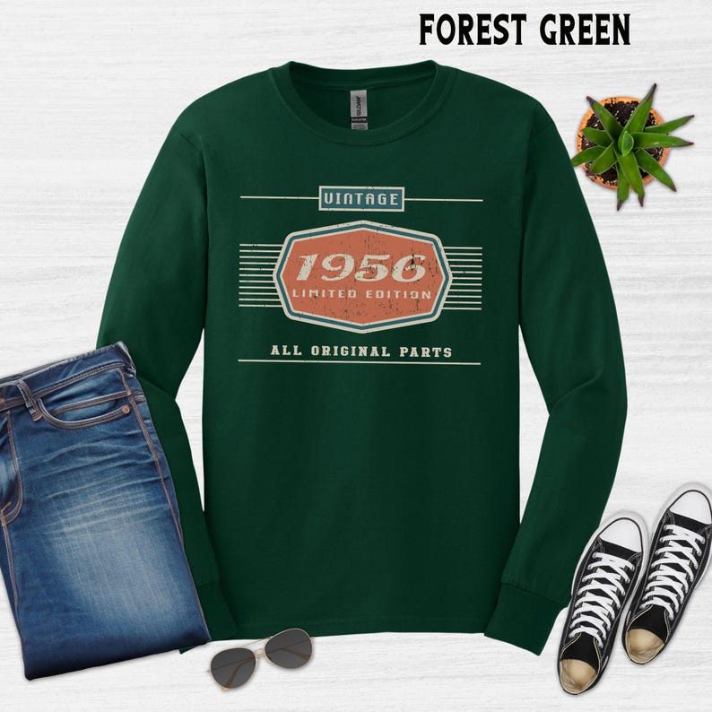 May include: Forest green long-sleeve shirt with a vintage design. The design includes the year "1956" in an orange and white hexagon, with the text "Limited Edition" and "All Original Parts". The shirt is paired with blue jeans and black and white sneakers.