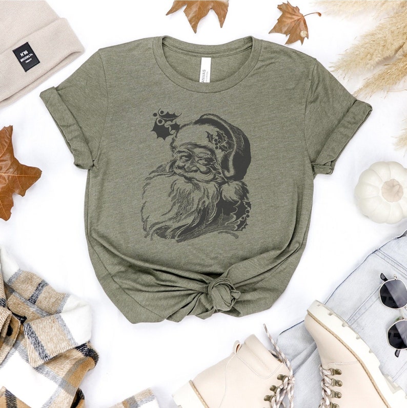 May include: A green t-shirt featuring a vintage illustration of Santa Claus on the front. The illustration is in black and white and shows Santa Claus with a beard and a hat.