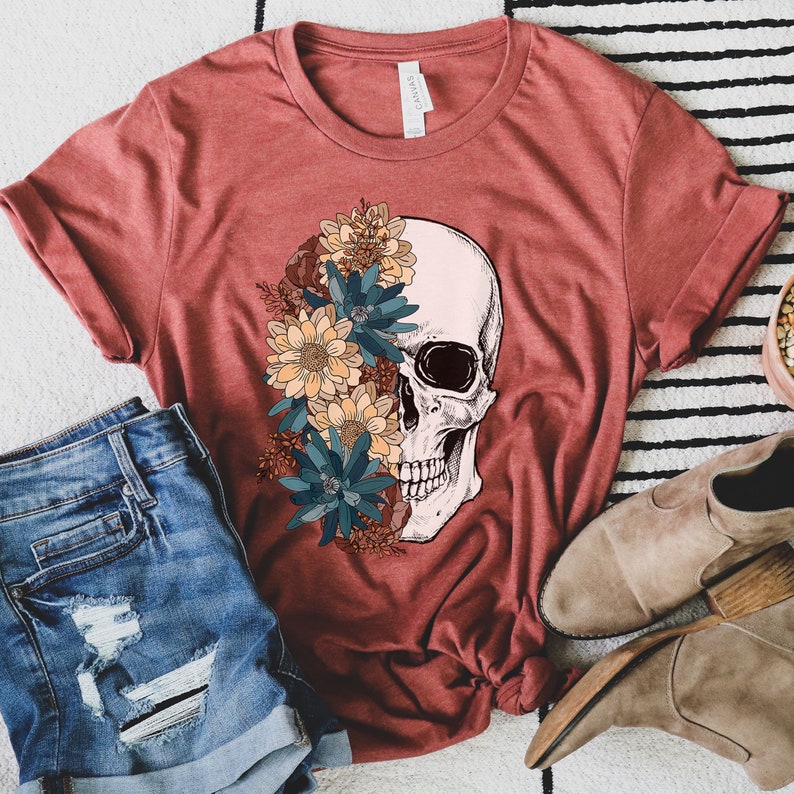 Op de afbeelding: Een rood T-shirt met een wit schedelontwerp. De schedel is half bedekt met een bloemstuk van blauwe, gele en oranje bloemen.