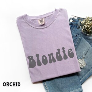 Puede incluir: Una camiseta de color lila claro con la palabra "Blondie" en letras grises. La camiseta est&aacute; colocada sobre unos vaqueros azules. La palabra "ORCHID" est&aacute; impresa en la esquina inferior izquierda.