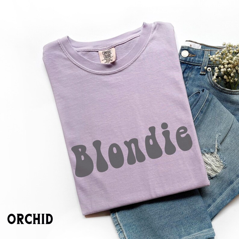 Puede incluir: Una camiseta de color lila claro con la palabra "Blondie" en letras grises. La camiseta est&aacute; colocada sobre unos vaqueros azules. La palabra "ORCHID" est&aacute; impresa en la esquina inferior izquierda.