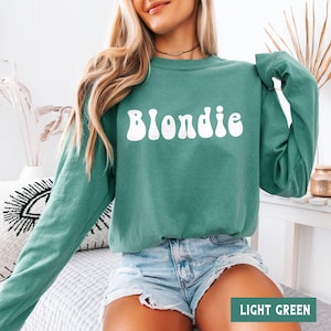Puede incluir: Una sudadera verde claro con la palabra "Blondie" en blanco, con letras de estilo retro. La sudadera tiene mangas largas y cuello redondo. La esquina inferior derecha de la imagen tiene el texto "LIGHT GREEN".