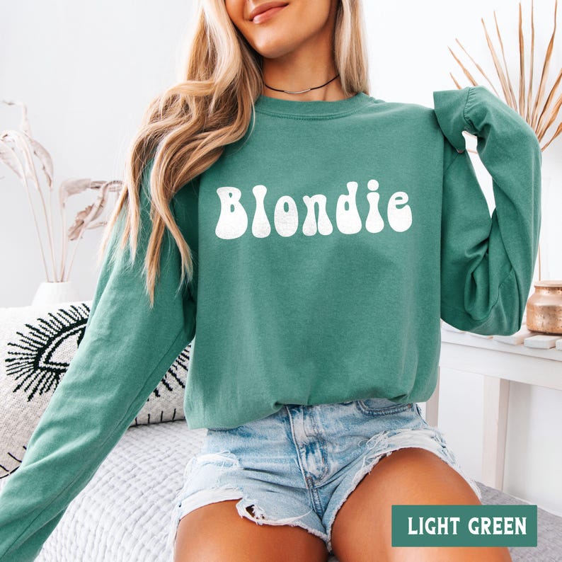 Puede incluir: Una sudadera verde claro con la palabra "Blondie" en blanco, con letras de estilo retro. La sudadera tiene mangas largas y cuello redondo. La esquina inferior derecha de la imagen tiene el texto "LIGHT GREEN".