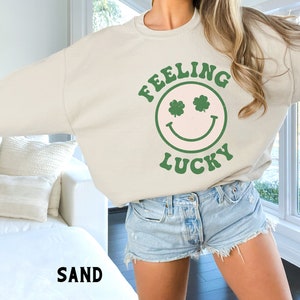 May include: A light beige sweatshirt with a green smiley face graphic that says "Feeling Lucky" with shamrock eyes. The model is wearing the sweatshirt with denim shorts.