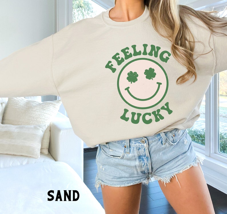 May include: A light beige sweatshirt with a green smiley face graphic that says "Feeling Lucky" with shamrock eyes. The model is wearing the sweatshirt with denim shorts.