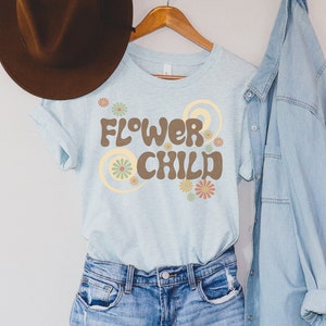 Puede incluir: Una camiseta azul claro con las palabras "FLOWER CHILD" en letras marrones, rodeada de dise&ntilde;os florales y espirales. La camiseta est&aacute; colgada en una percha de madera con un sombrero marr&oacute;n y una camisa vaquera en el fondo.