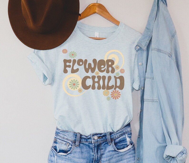 Puede incluir: Una camiseta azul claro con las palabras "FLOWER CHILD" en letras marrones, rodeada de dise&ntilde;os florales y espirales. La camiseta est&aacute; colgada en una percha de madera con un sombrero marr&oacute;n y una camisa vaquera en el fondo.