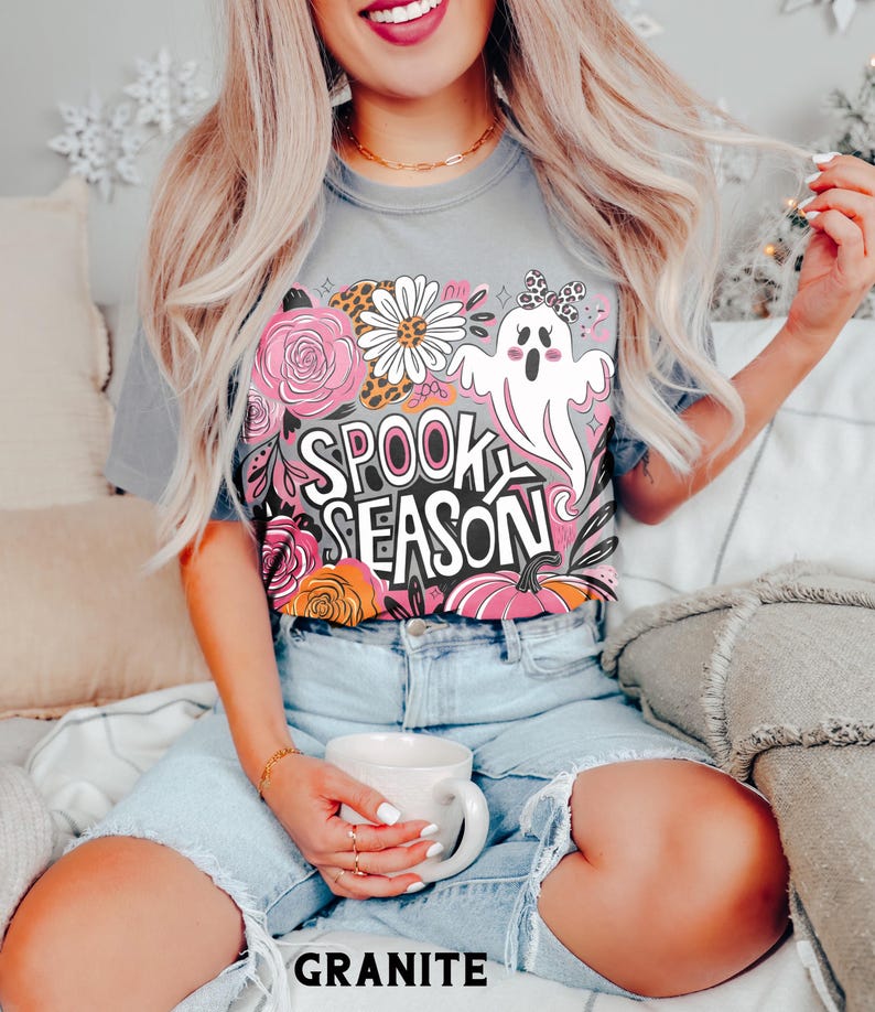 May include: A gray t-shirt with a colorful "Spook Season" graphic featuring a ghost, flowers, and pumpkins. The word "GRANITE" is printed at the bottom. The person is wearing denim shorts and holding a white mug.