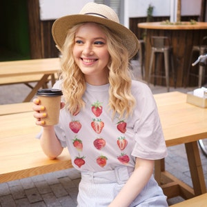 Op de afbeelding: Een wit T-shirt met een patroon van rode aardbeien. De persoon die het shirt draagt, houdt een bruin papieren bekertje vast.