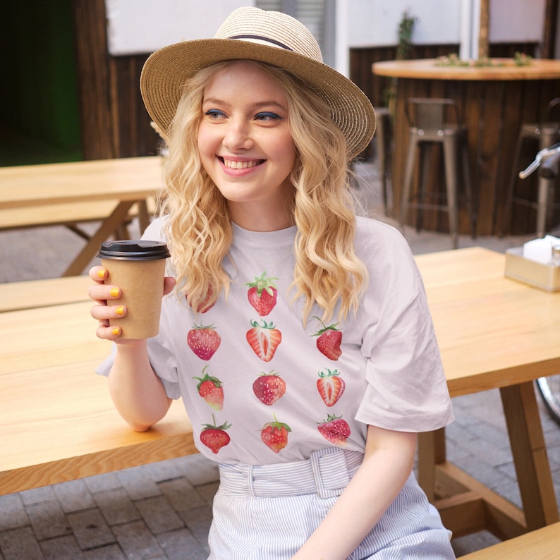 Op de afbeelding: Een wit T-shirt met een patroon van rode aardbeien. De persoon die het shirt draagt, houdt een bruin papieren bekertje vast.