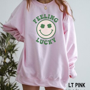 May include: A light pink sweatshirt with a green smiley face graphic that says "Feeling Lucky" on it. The model is wearing a size large.