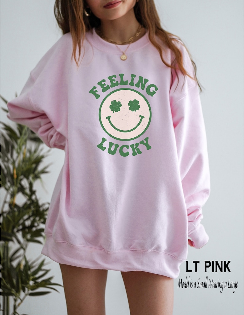 May include: A light pink sweatshirt with a green smiley face graphic that says "Feeling Lucky" on it. The model is wearing a size large.