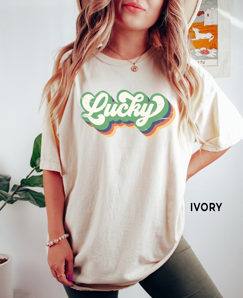May include: A cream colored t-shirt with a retro-style graphic that says "Lucky" in green, yellow, orange, and blue. The text is in a faded, vintage style. The shirt is shown on a model wearing it. The text "IVORY" is visible in the bottom right corner of the image.