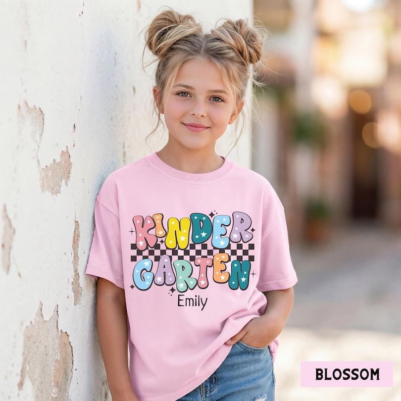May include: A light pink t-shirt with the words "Kindergarten" in colorful bubble letters and a checkered stripe. The name "Emily" is printed below the word "Kindergarten". The shirt is worn by a young girl.