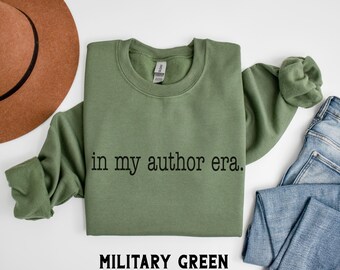 May include: A military green sweatshirt with the phrase "in my author era." printed in black. The words "MILITARY GREEN" are printed in a bold, sans-serif font below the sweatshirt. A brown hat and blue jeans are also visible.