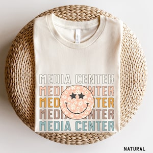 Op de afbeelding: Een beige T-shirt met de tekst "MEDIA CENTER" die in een gestapelde patroon wordt herhaald met een glimlachend gezicht met een madeliefjespatroon in het midden. De tekst is in verschillende kleuren, waaronder oranje, geel, blauw en bruin. Het T-shirt ligt op een geweven strooppervlak. Het woord "NATURAL" is in zwart onderaan de afbeelding gedrukt.