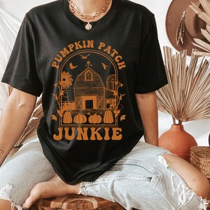 Puede incluir: Camiseta negra con un gr&aacute;fico naranja de un granero, un molino de viento y calabazas con el texto "Pumpkin Patch Junkie" alrededor del gr&aacute;fico.