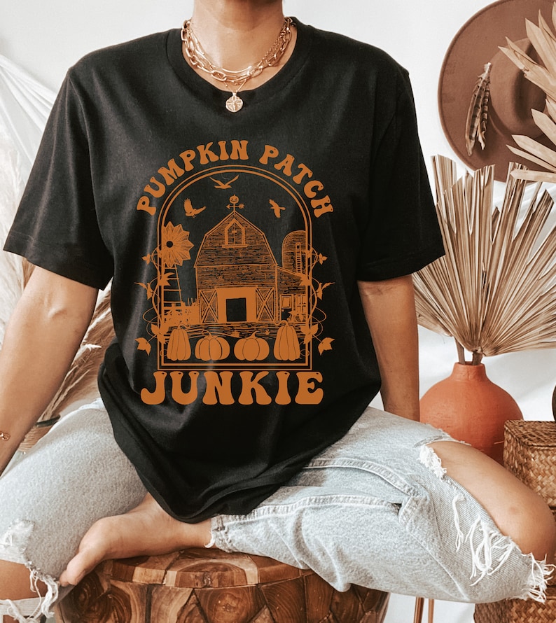 Puede incluir: Camiseta negra con un gr&aacute;fico naranja de un granero, un molino de viento y calabazas con el texto "Pumpkin Patch Junkie" alrededor del gr&aacute;fico.