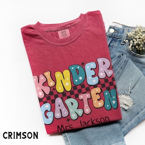 May include: Crimson-colored t-shirt with the words "KINDER GARDEN" in colorful, bubble letters with star accents. The shirt is paired with denim shorts and a sprig of white flowers. The text "Mrs. Jackson" is below the main text.