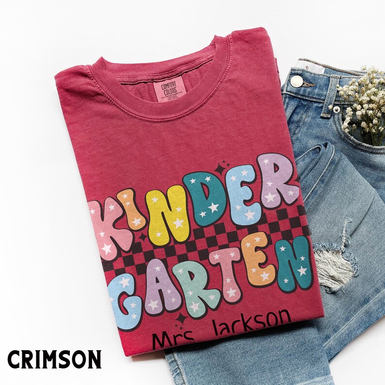 May include: Crimson-colored t-shirt with the words "KINDER GARDEN" in colorful, bubble letters with star accents. The shirt is paired with denim shorts and a sprig of white flowers. The text "Mrs. Jackson" is below the main text.