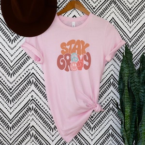 May include: Pink t-shirt with the text "Stay Groovy" in a retro font with peace signs and smiley faces.