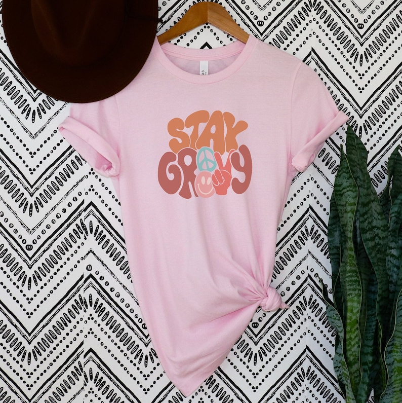 May include: Pink t-shirt with the text "Stay Groovy" in a retro font with peace signs and smiley faces.