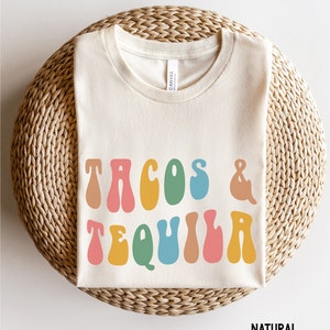 Puede incluir: Una camiseta de color natural con el texto "TACOS & TEQUILA" en una fuente retro con letras de colores.