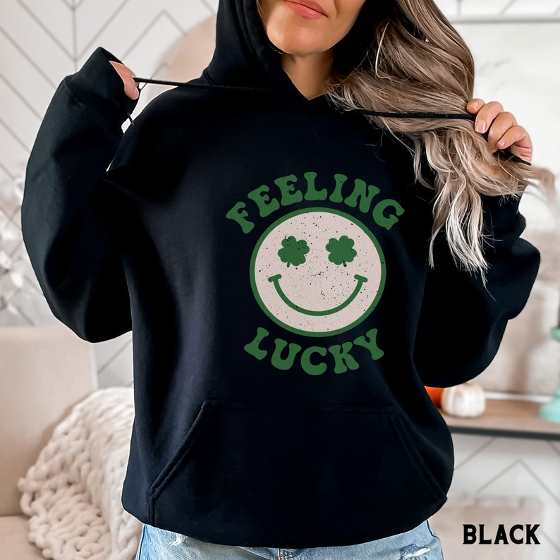May include: Black hoodie with a white smiley face with green shamrocks for eyes and the text "Feeling Lucky" in a retro font.