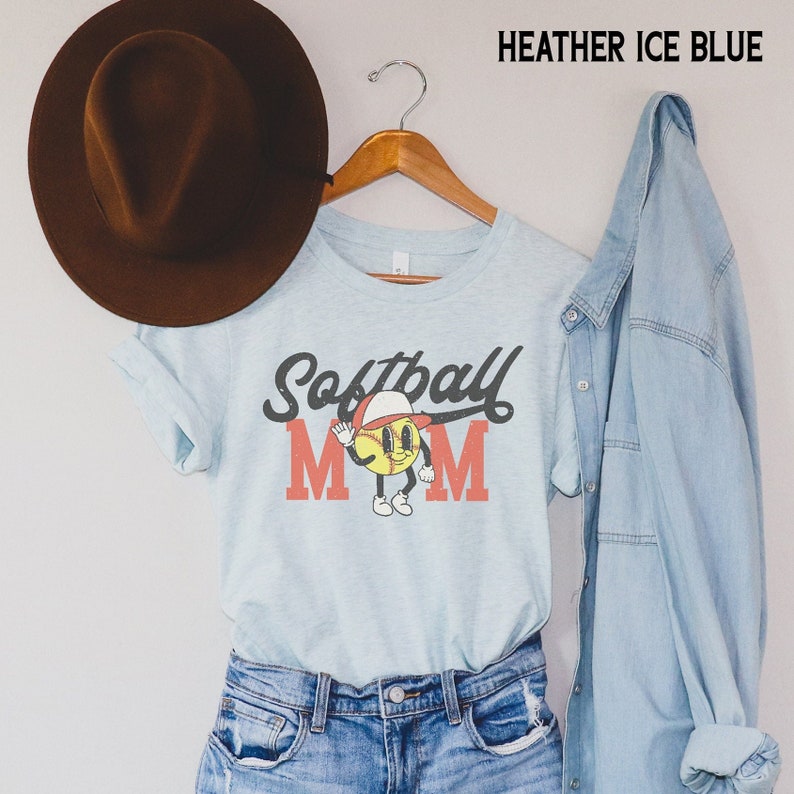 May include: Heather ice blue t-shirt with the words "Softball Mom" in a vintage style. The "O" in Mom is replaced with a cartoon softball character wearing a baseball cap. A brown hat and denim jacket are also in the image.