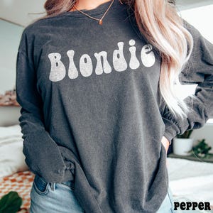 Puede incluir: Una camiseta de manga larga gris oscuro con la palabra "Blondie" en blanco, con letras de estilo retro. La camiseta se combina con vaqueros azules. La palabra "pepper" est&aacute; impresa en la esquina inferior derecha.