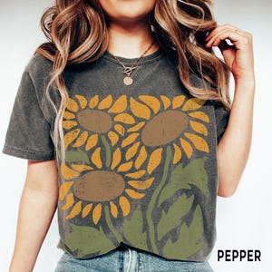 May include: A dark gray t-shirt with a vintage-style graphic of three sunflowers in shades of yellow, brown, and green. The text "PEPPER" is visible in the bottom right corner of the image.