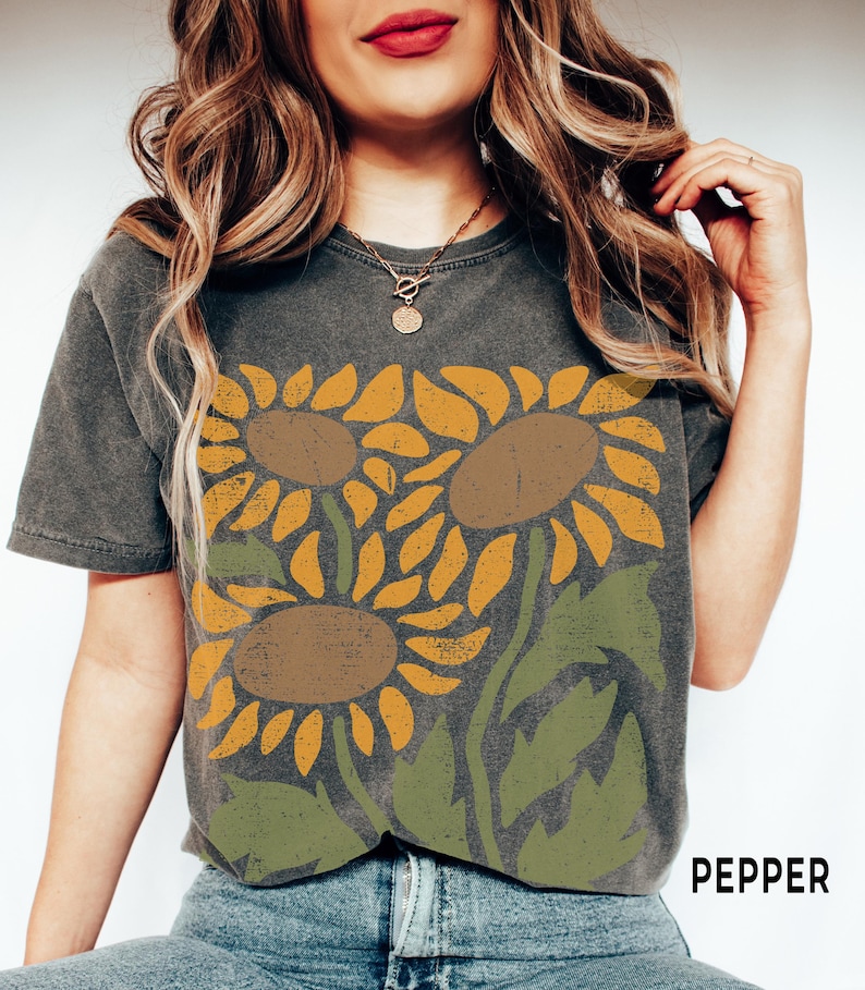 May include: A dark gray t-shirt with a vintage-style graphic of three sunflowers in shades of yellow, brown, and green. The text "PEPPER" is visible in the bottom right corner of the image.