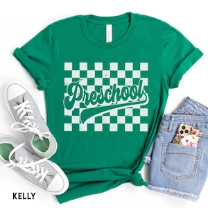 Pu&ograve; includere: Una maglietta verde con la scritta "Preschool" in bianco, con un font retr&ograve;, su uno sfondo a scacchi. La maglietta &egrave; abbinata a scarpe da ginnastica grigie e pantaloncini di jeans. La parola "KELLY" &egrave; stampata in basso.
