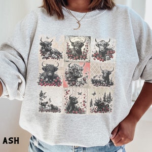 May include: A gray sweatshirt with a patchwork design featuring black and white illustrations of Highland cattle with red berries and butterflies. The text "ASH" is printed on the bottom left corner of the image.