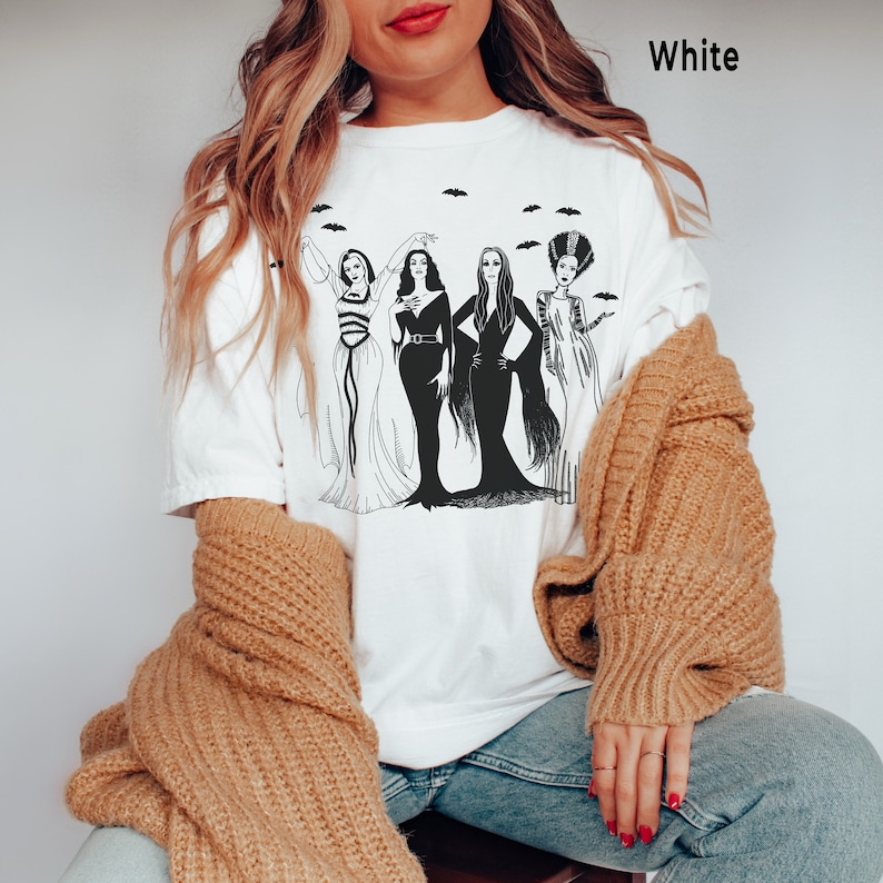 K&ouml;nnte beinhalten: Wei&szlig;es T-Shirt mit einer Schwarzwei&szlig;illustration von vier Frauen in Halloweenkost&uuml;men. Die Frauen sind als Vampir, Hexe, Geist und Zombie verkleidet. Der Text "White" ist in der oberen rechten Ecke des Bildes sichtbar.