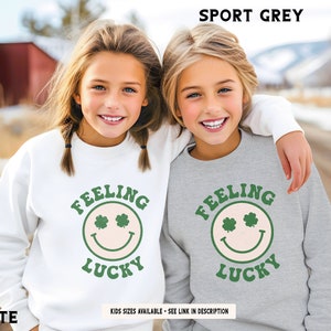 May include: Two children wearing matching sweatshirts with a green shamrock smiley face and the text "Feeling Lucky". One sweatshirt is white and the other is sport grey.