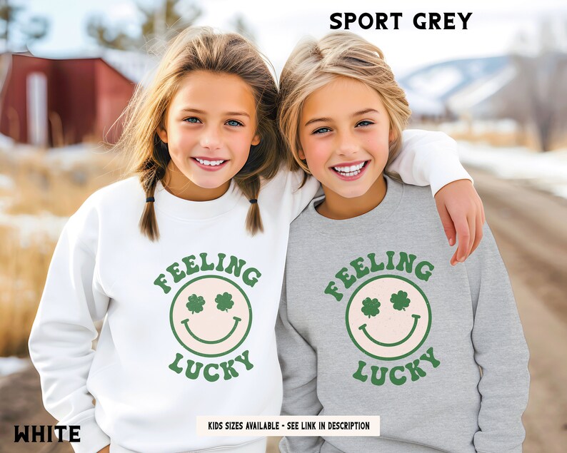 May include: Two children wearing matching sweatshirts with a green shamrock smiley face and the text "Feeling Lucky". One sweatshirt is white and the other is sport grey.